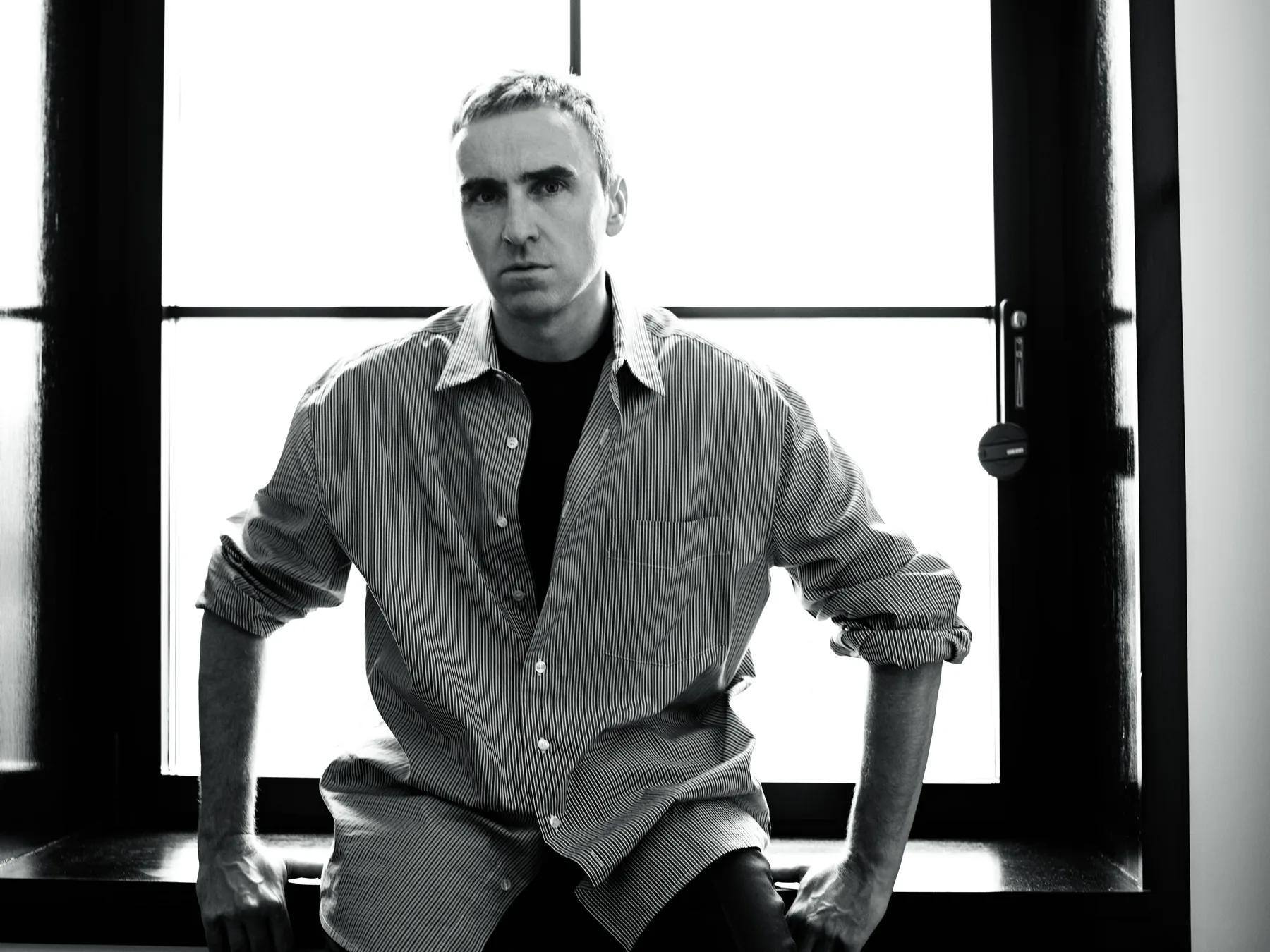 Raf Simons seated on a window sill in a striped shirt over a black T-shirt, black-and-white portrait