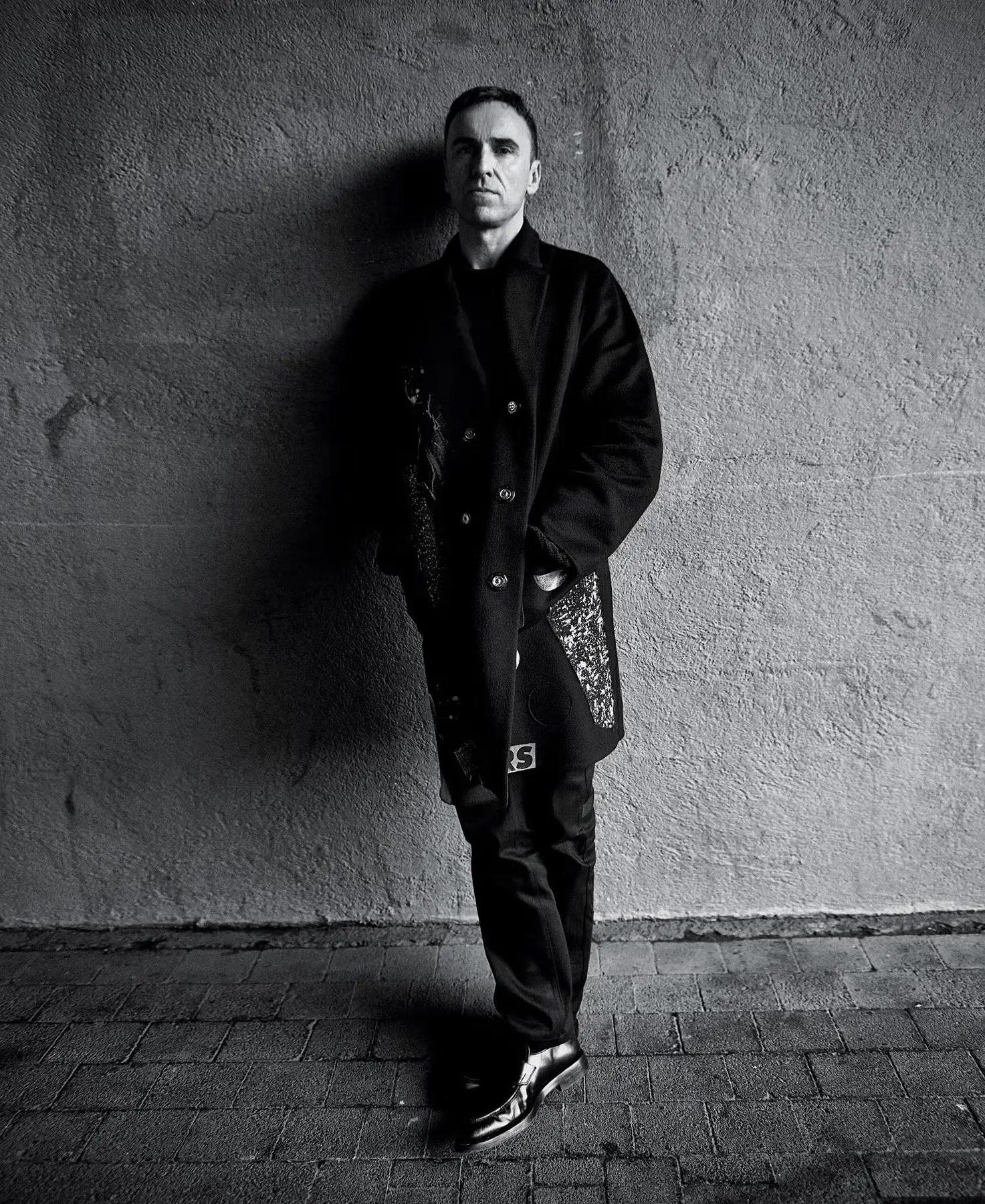 Raf Simons standing against a wall in a long black coat, black-and-white portrait