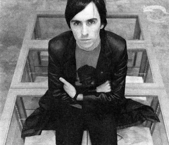 Raf Simons in the 1990s in a leather coat, seated on a wooden structure, black-and-white portrait