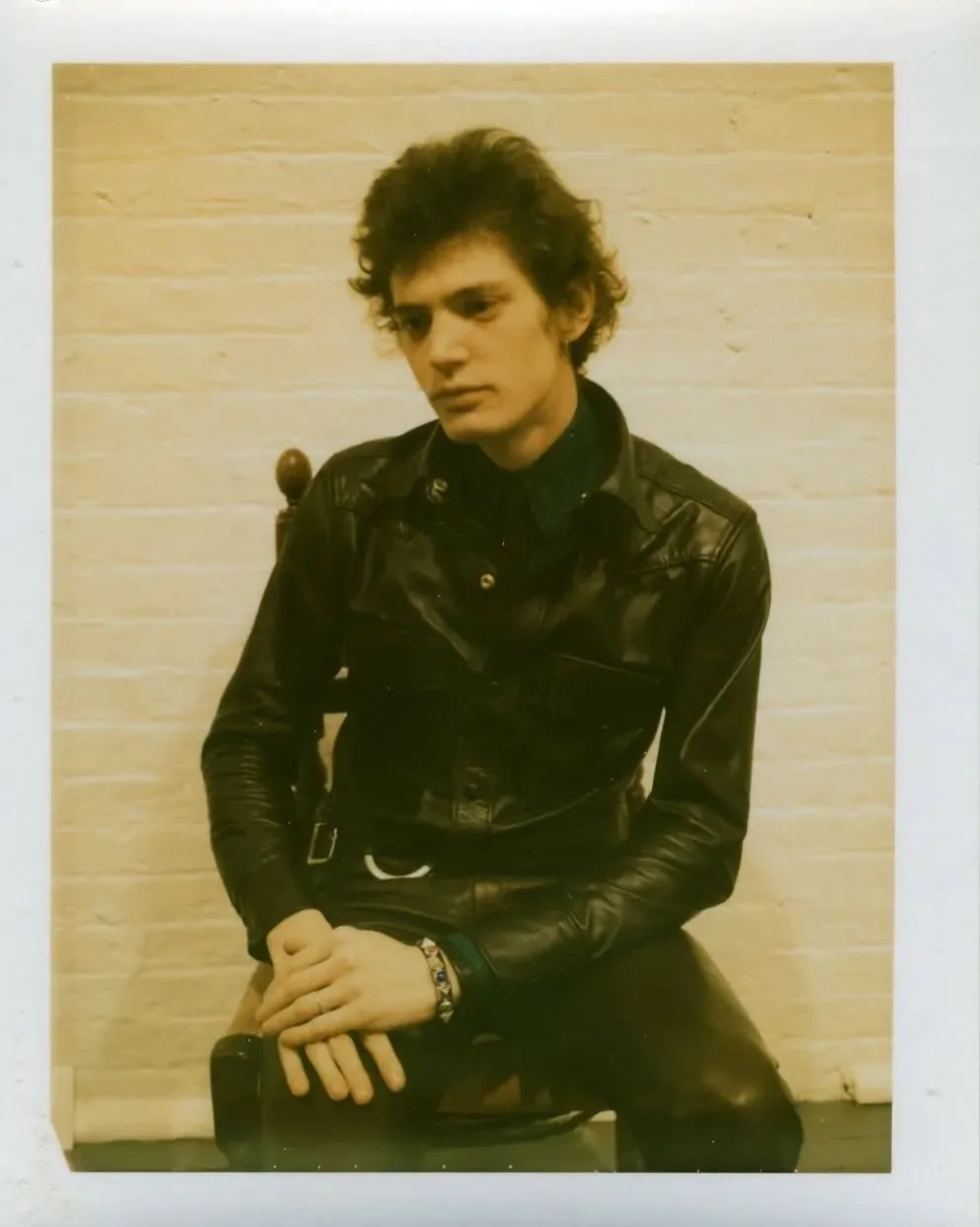 Robert Mapplethorpe in black leather, seated against a white brick wall, Polaroid