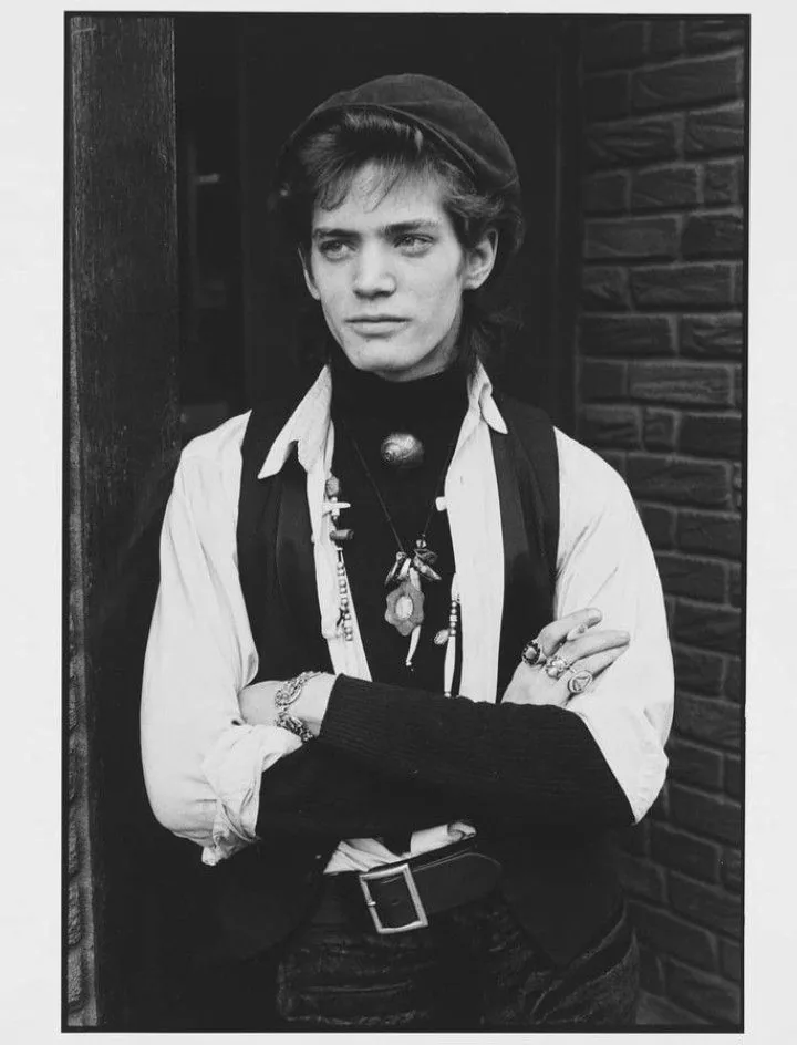 Robert Mapplethorpe with arms crossed, cap, necklaces and rings, against a brick wall, black-and-white