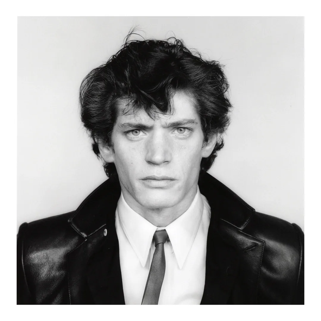 Robert Mapplethorpe in a leather jacket, white shirt and thin tie, black-and-white portrait