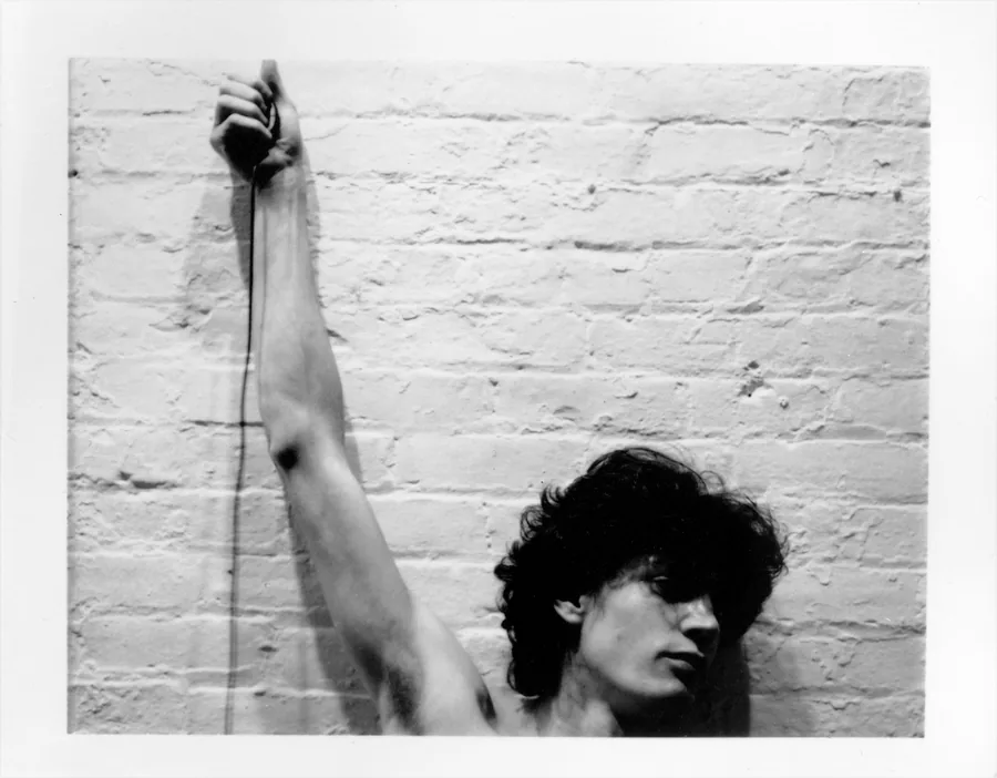 Robert Mapplethorpe with one arm raised against a white brick wall, black-and-white Polaroid