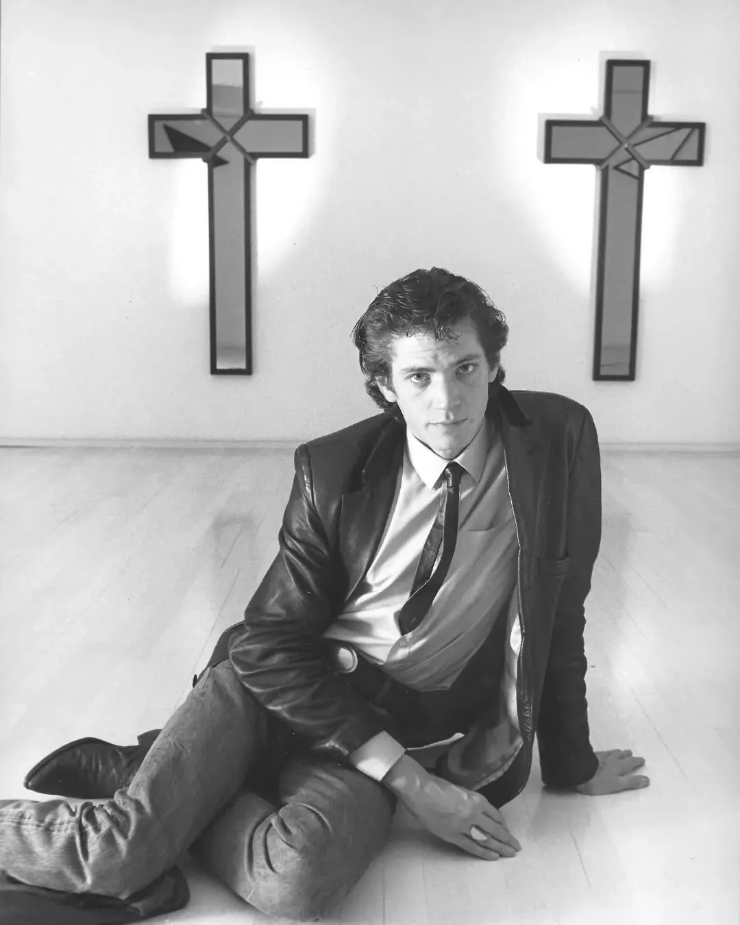 Robert Mapplethorpe seated on a wooden floor in front of two mirrored crosses, black-and-white